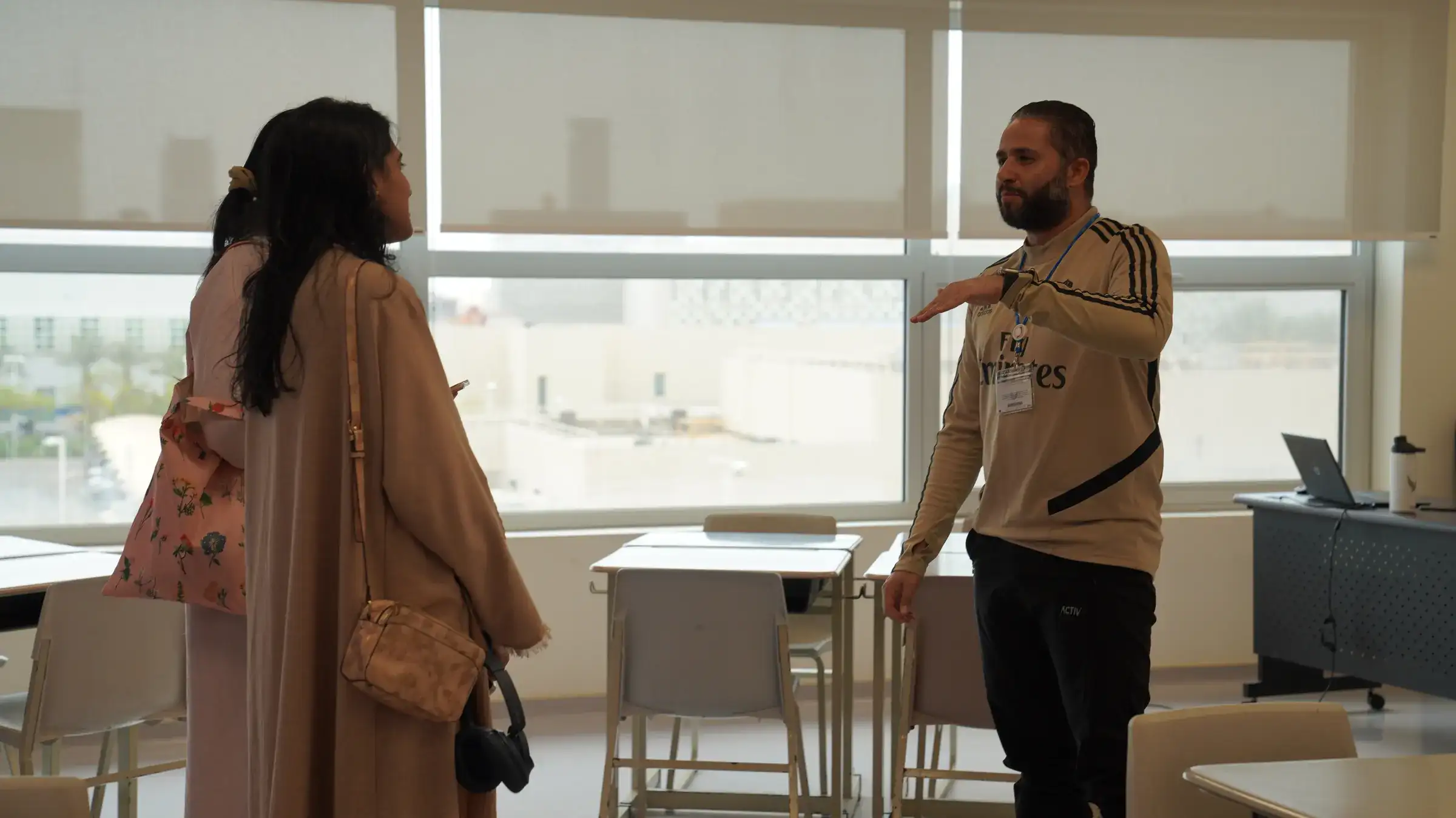 أحد أعضاء طاقم AISJ يرتدي قميصاً يحمل شعار المدرسة ويشير نحو المكان بينما يتحدث مع سيدة ترتدي الحجاب والعباءة في فضاء مضيء تحيط به النوافذ.