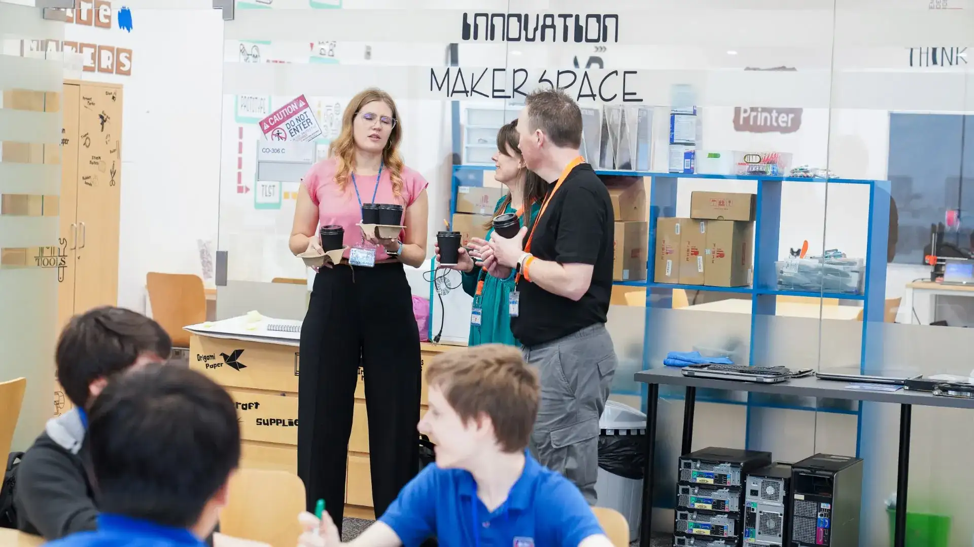 Staff speak with students inside the Innovation MakerSpace, with a Printer sign, craft-supply cabinets and computer towers visible behind the glass.