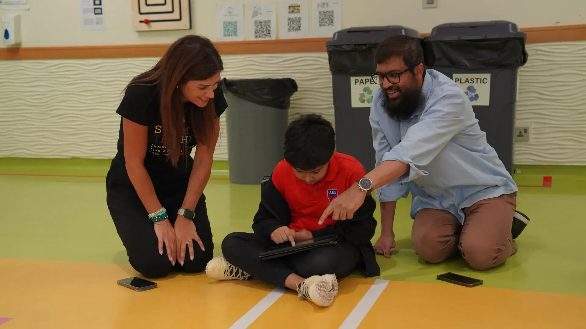 Two teachers kneel on a gym floor beside an elementary student, one pointing at the tablet he is using.
