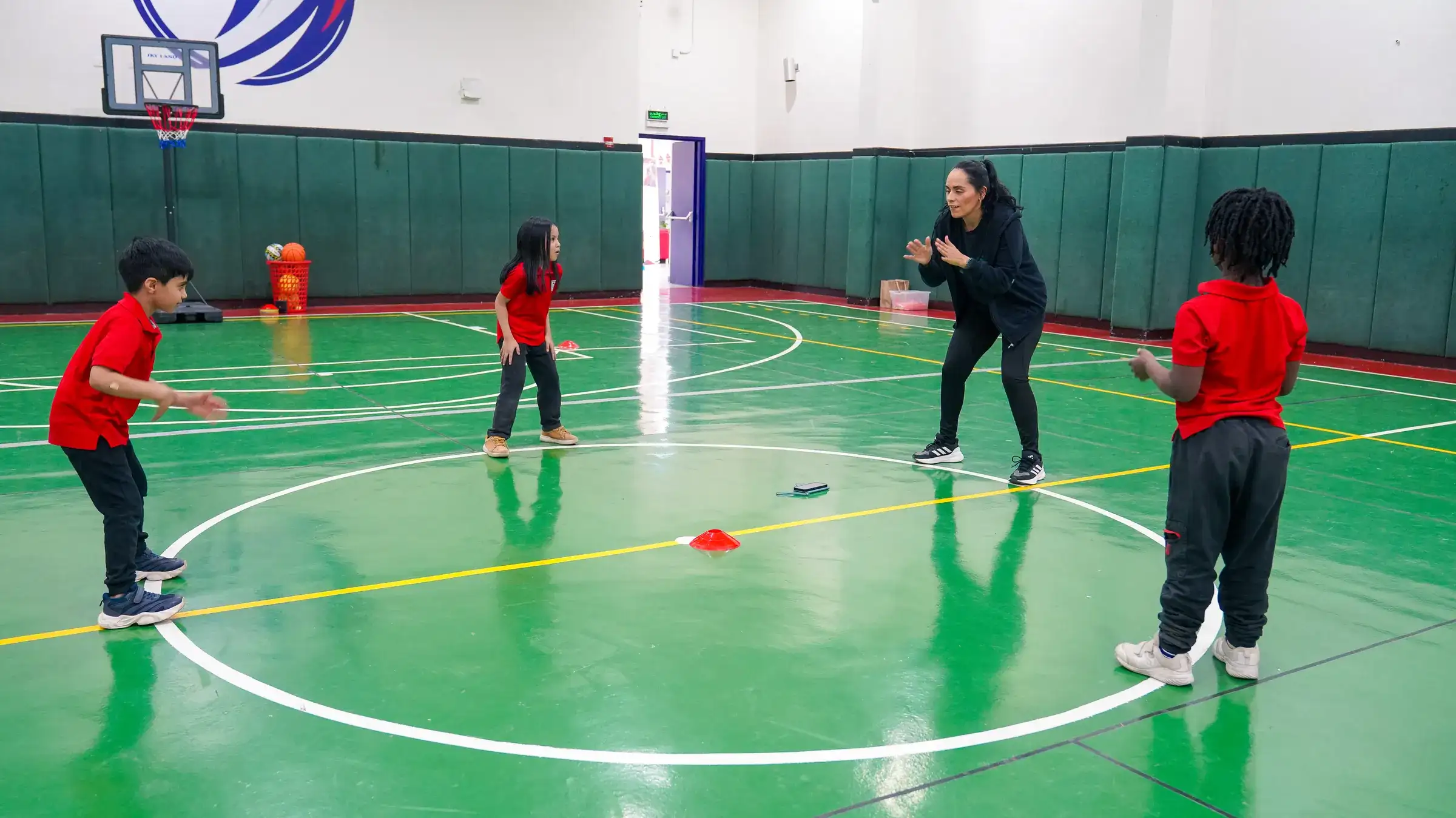 A PE teacher and elementary students play a ball game on the marked green court of the AISJ gymnasium, with banners visible on the walls.