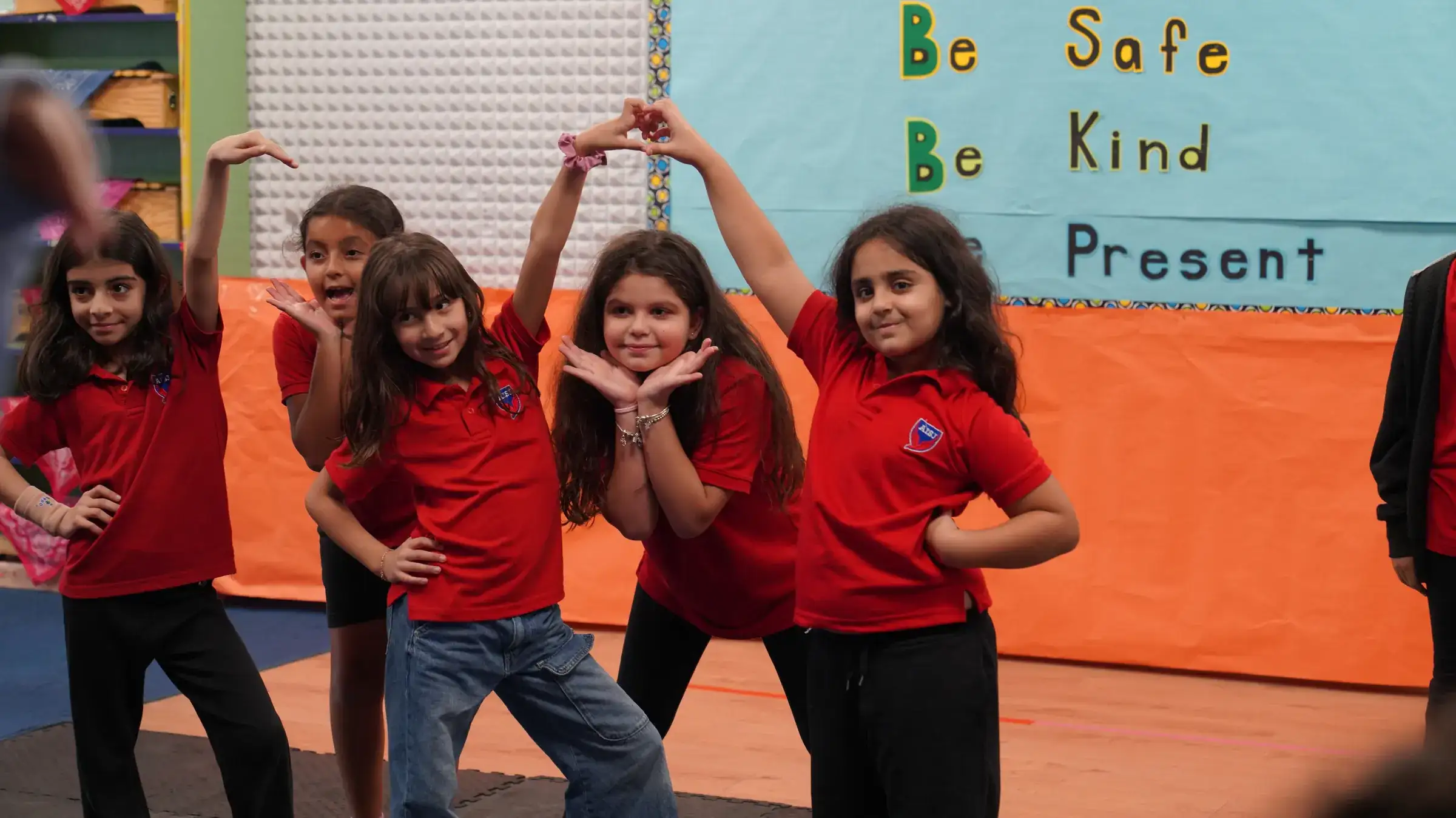 Five elementary students in red AISJ polo shirts pose together in front of a classroom sign reading 'Be Safe, Be Kind, Be Present.'