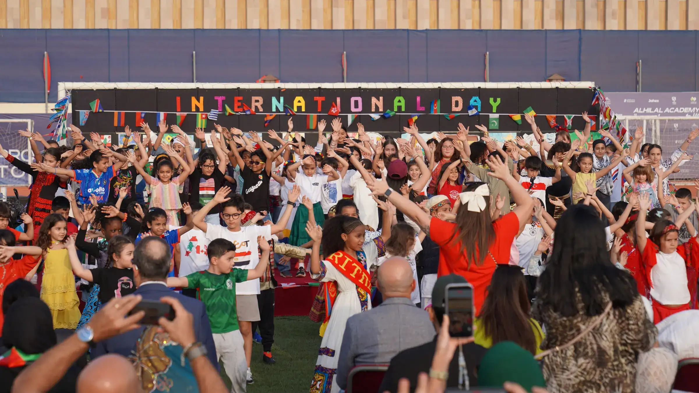 Students dance together on a sports field beneath a colorful 'International Day' sign strung with national flags.