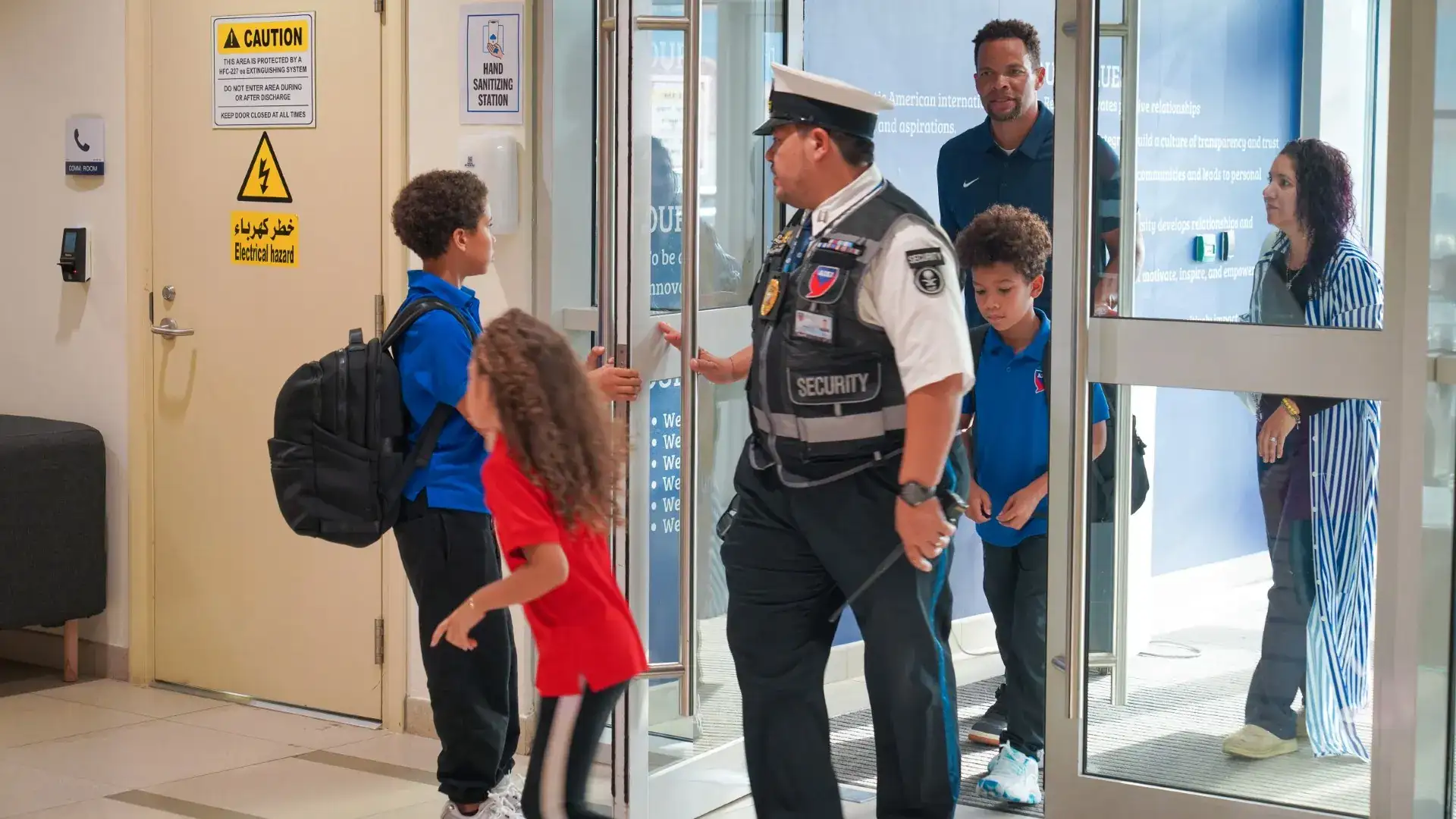 A uniformed security officer walks two students in AISJ uniforms through a glass entrance door, with an adult and another staff member behind them.