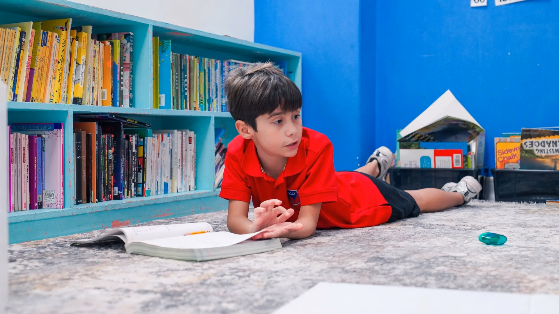 A young AISJ student lying on the library floor reading a book beside the shelves