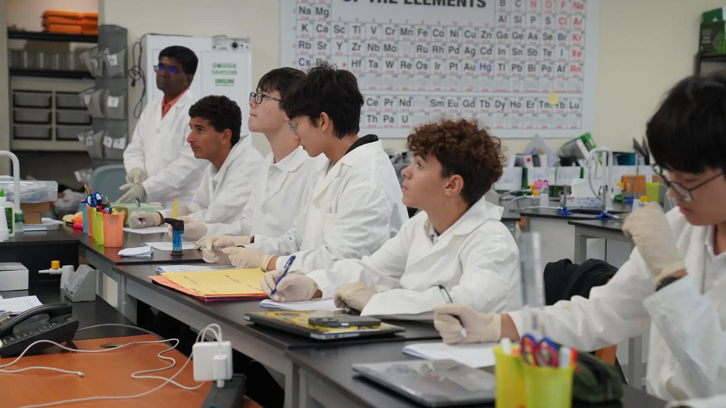 High school students in lab coats and gloves take notes at a lab bench while their teacher stands behind them, beneath a large periodic table.