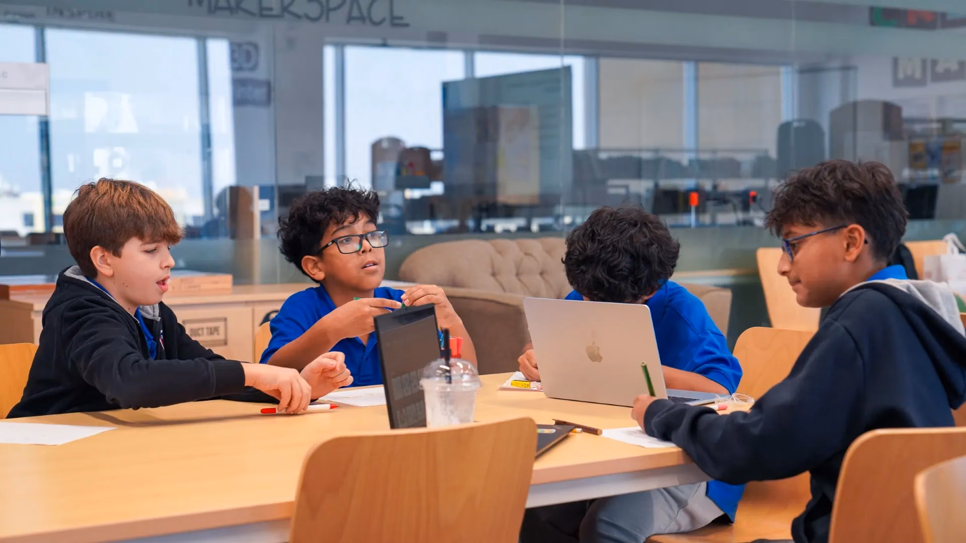 Four students talk through a task at a round table with a laptop and a tablet, with MAKERSPACE lettering on the glass wall behind them.