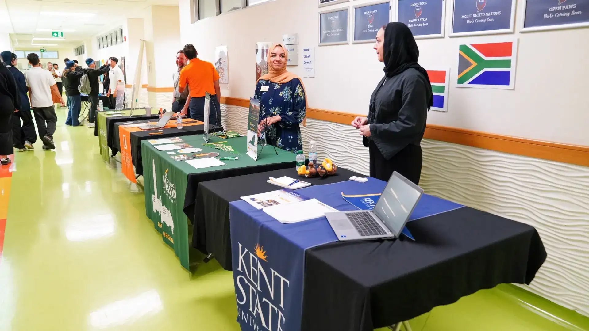 Students and staff talk with university representatives at booths for George Mason, Kent State and other universities in a school hallway.