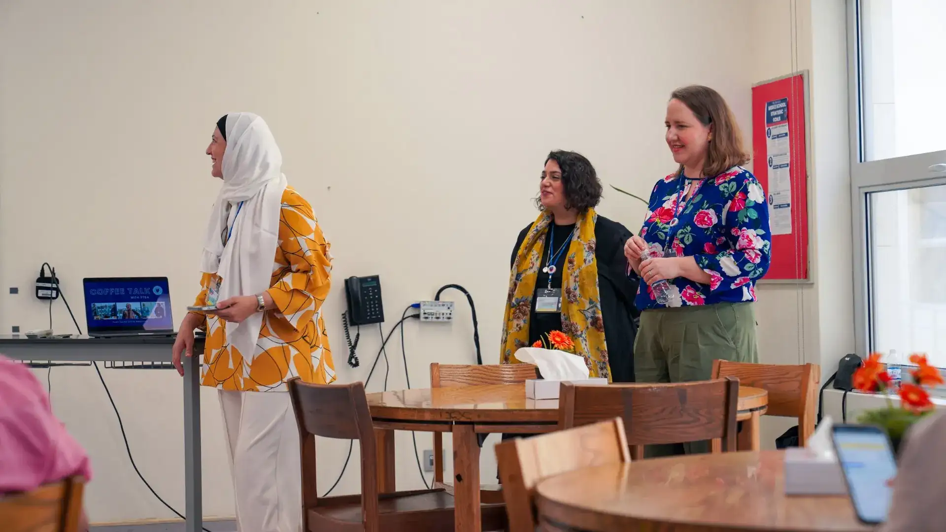 Three women stand and talk in a room set up for a school event, with a laptop on a table beside them displaying 'Coffee Talk with PTSA.'