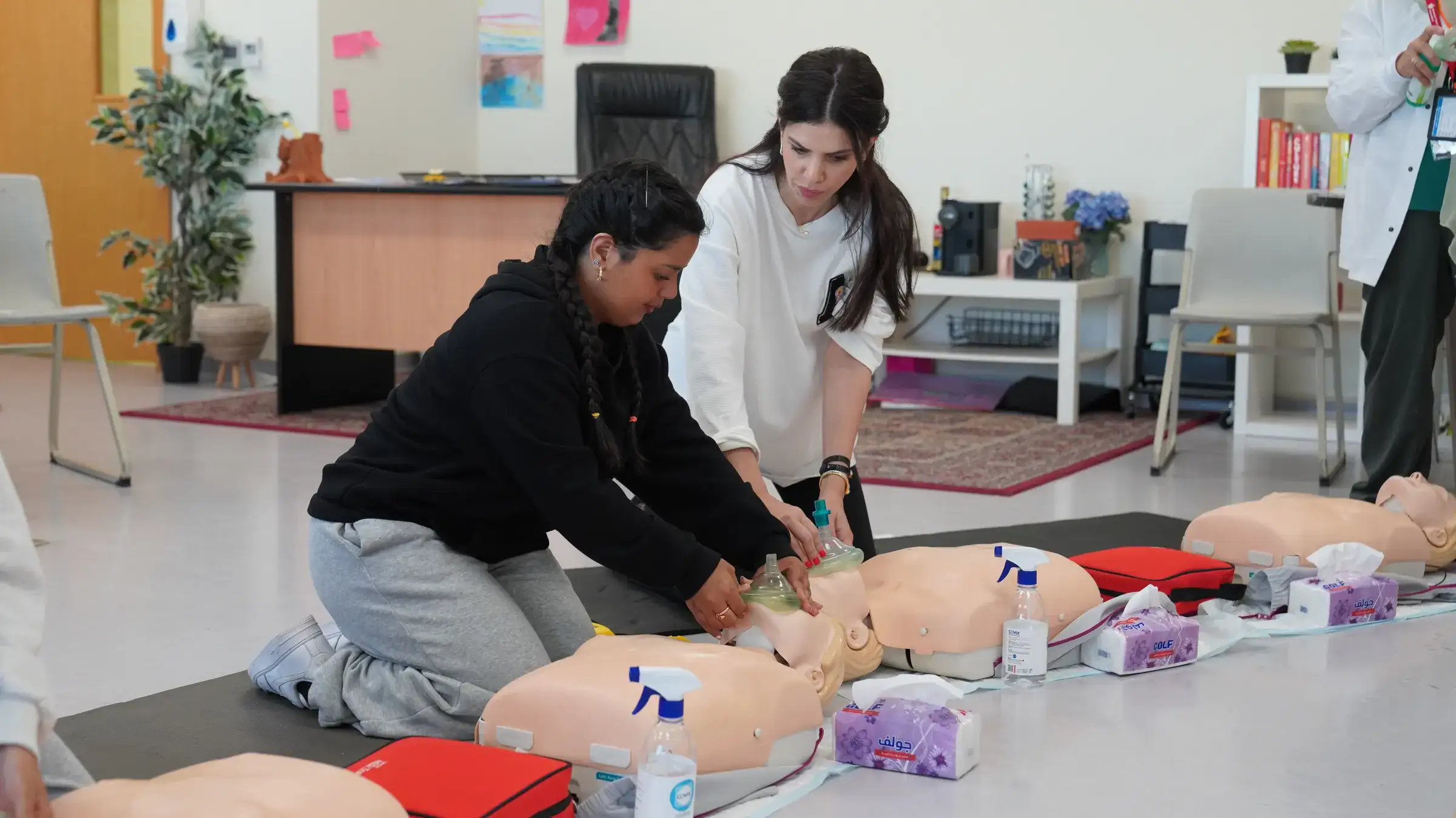 Two AISJ students kneel and practice hands-on CPR technique on medical training mannequins while a staff member guides them in a bright classroom.