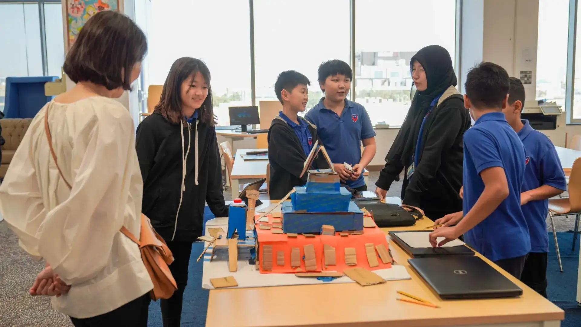 Middle school students and visitors gather around a cardboard-and-paper model on a table beside two laptops.